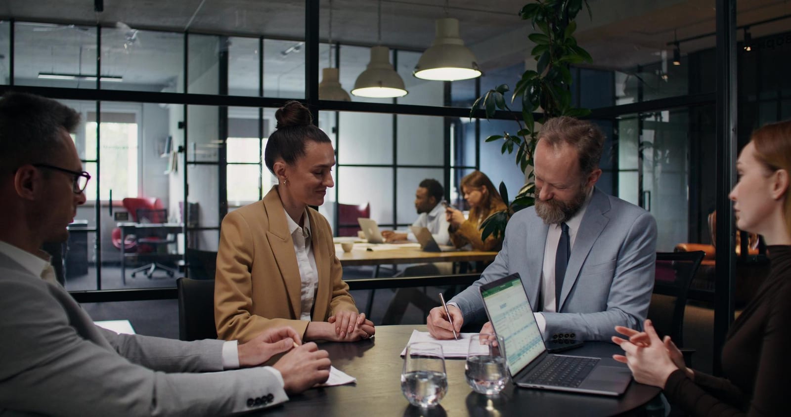 Vier Personen besprechen sich an einem runden Tisch in einem Büro mit Glaswänden, Laptop und Unterlagen.